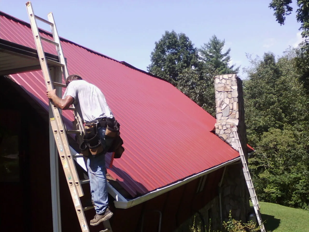 Roofer on a ladder beside a red metal roof and stone chimney — Soffit & Fascia Repair by Roofing Brothers in Brockton